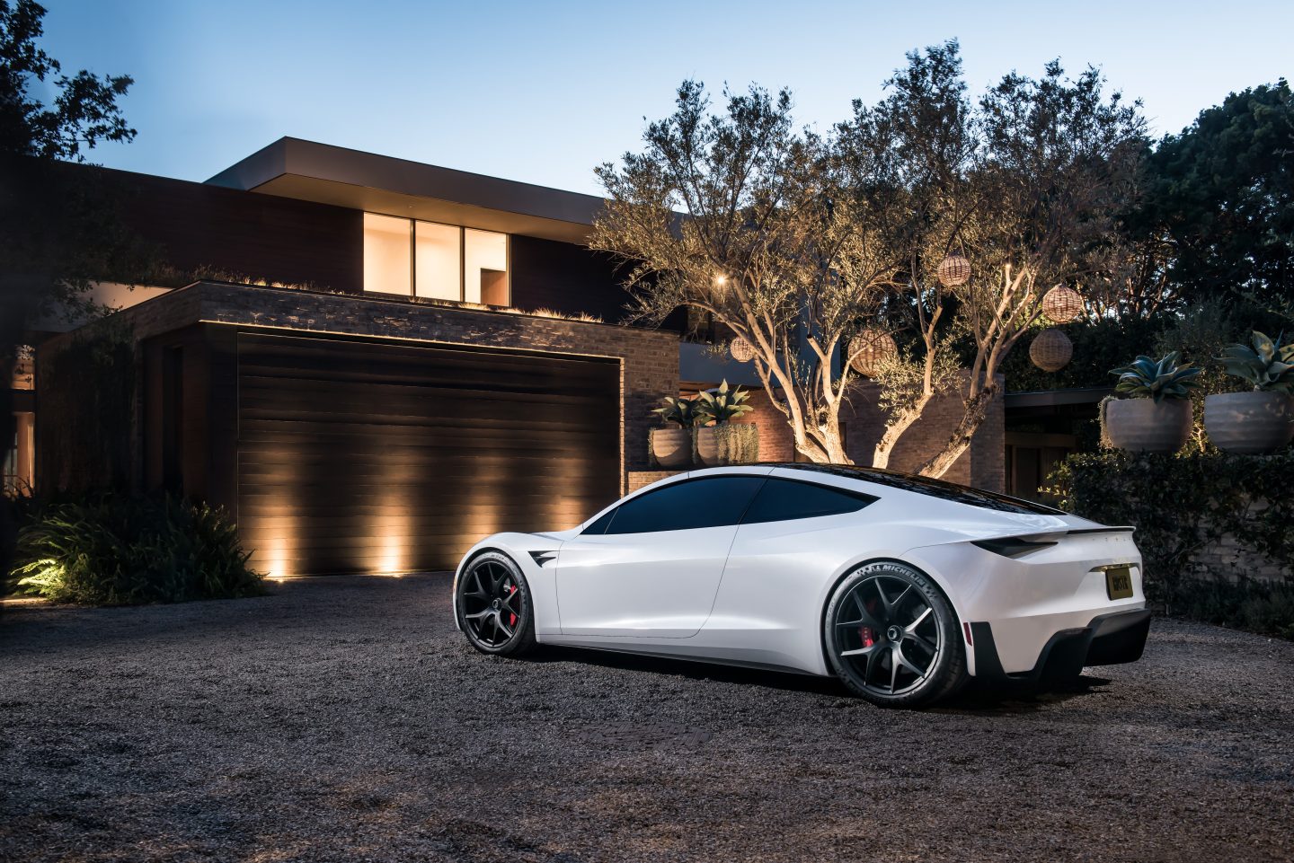 En hvit Tesla Roadster står i en gruset oppkjørsel foran et moderne hus